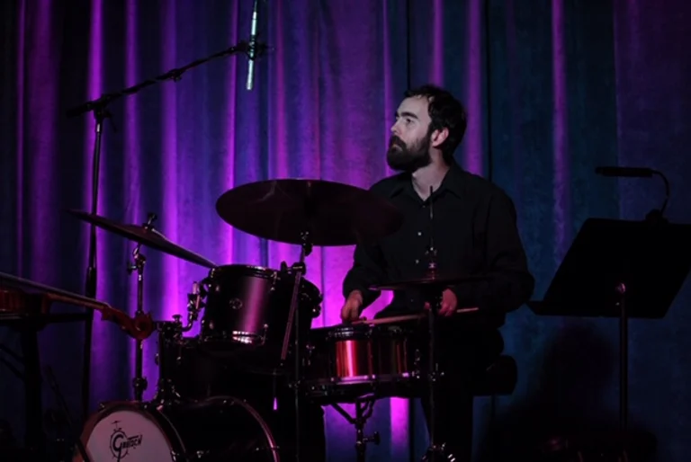 Dark-haired man in black button-up shirt sits at a drum set on stage in front of lighted, purple curtains. He holds sticks in his hands as he plays the snare drum, looking off to our left in concentration.
