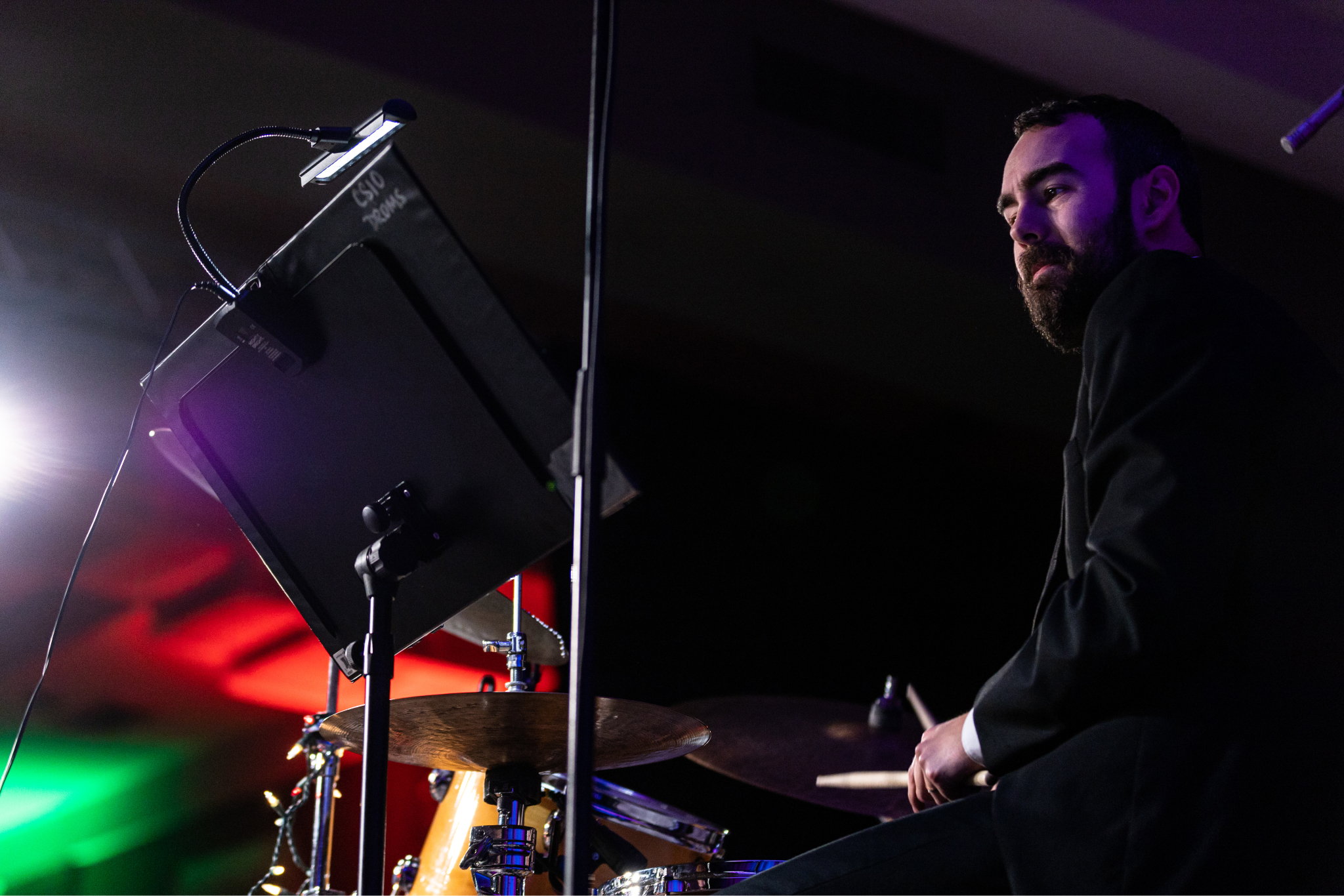 Man in dark suit sits at a drum set, drumsticks in hand, looking to a sheet stand of music on his left in concentration.